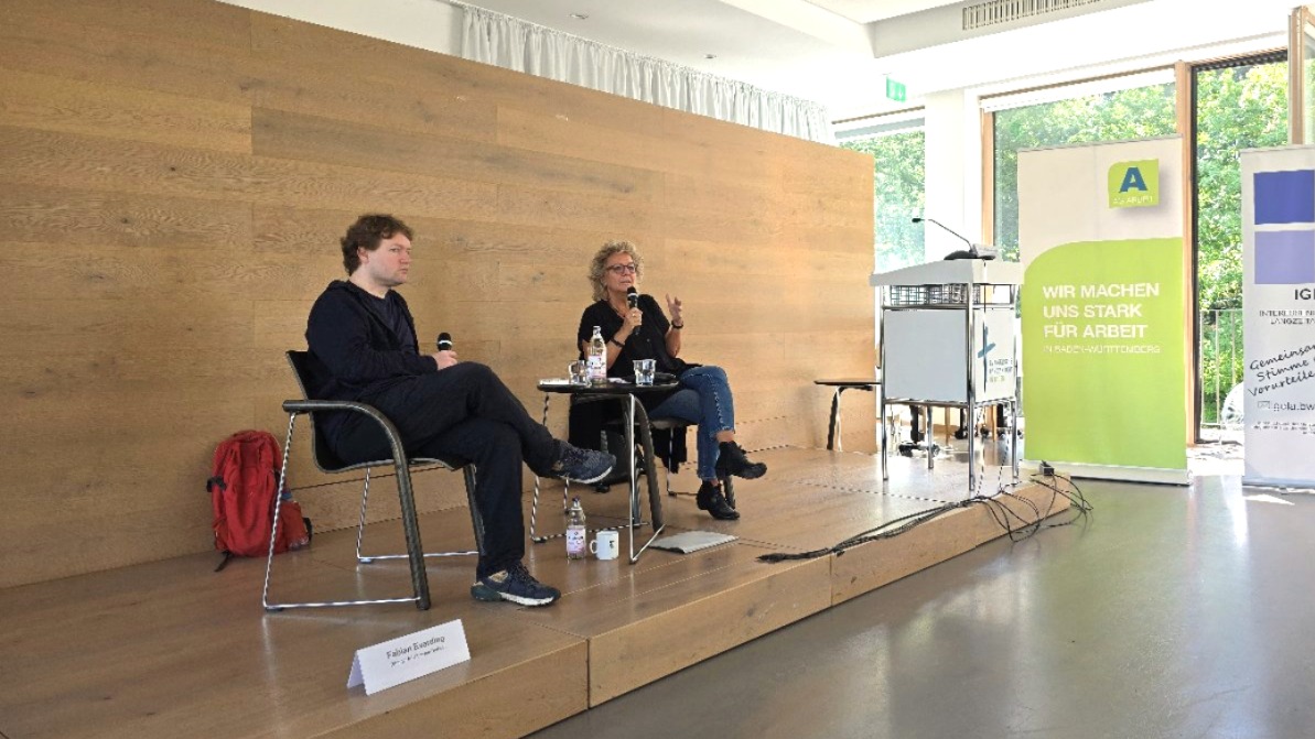 Beate Müller-Gemmeke sitzt mit einem Gesprächspartner auf einer kleinen Bühne, beide halten Mikrofone in der Hand. Im Hintergrund eine schlichte Holzverkleidung. Auf dem Tisch stehen Wasserflaschen und Tassen – eine ruhige Gesprächssituation im Rahmen der Arbeitslosentagung.