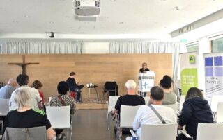 Beate Müller-Gemmeke spricht am Rednerpult vor dem Publikum der Arbeitslosentagung. Im Hintergrund sind Roll-ups der Veranstalter zu sehen, vor ihr sitzen aufmerksam zuhörende Teilnehmende in hellen Stuhlreihen.