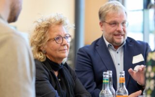 Beate Müller-Gemmeke steht mit anderen Teilnehmenden an einem Tisch beim Fachtag in Berlin. Neben ihr spricht ein Mann im blauen Sakko. Vor ihnen stehen Getränkeﬂaschen. Beide wirken konzentriert im Gespräch.