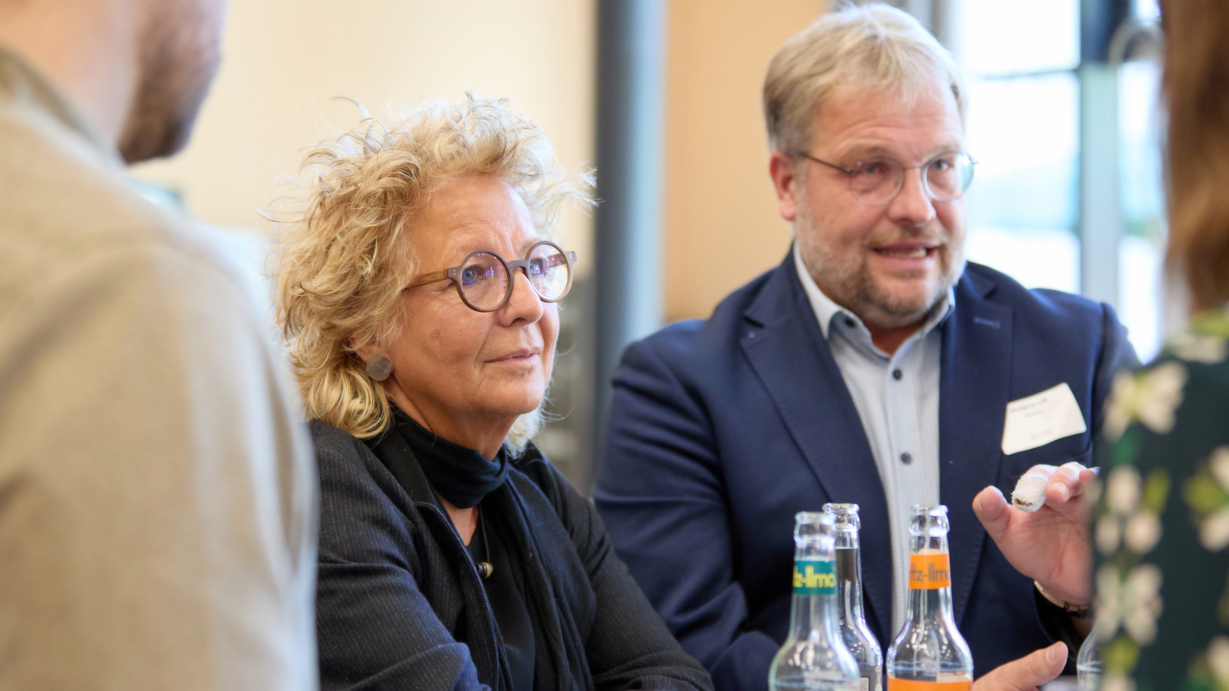 Beate Müller-Gemmeke steht mit anderen Teilnehmenden an einem Tisch beim Fachtag in Berlin. Neben ihr spricht ein Mann im blauen Sakko. Vor ihnen stehen Getränkeflaschen. Beide wirken konzentriert im Gespräch.