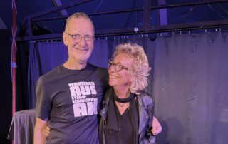 Beate Müller-Gemmeke steht lächelnd neben einem Bekannten bei einer Geburtstagsfeier. Beide schauen sich herzlich an und strahlen Freude und Vertrautheit aus. Im Hintergrund eine Bühne mit Vorhang – die Atmosphäre wirkt lebendig und warm.