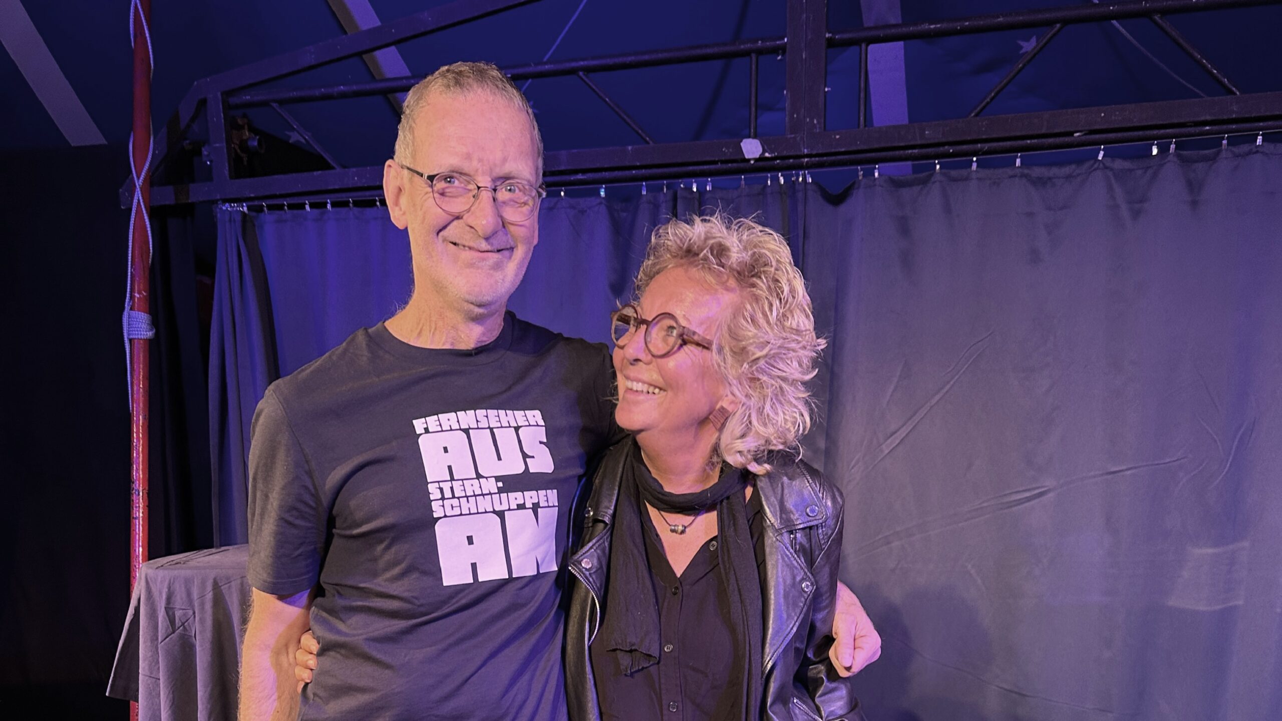 Beate Müller-Gemmeke steht lächelnd neben einem Bekannten bei einer Geburtstagsfeier. Beide schauen sich herzlich an und strahlen Freude und Vertrautheit aus. Im Hintergrund eine Bühne mit Vorhang – die Atmosphäre wirkt lebendig und warm.