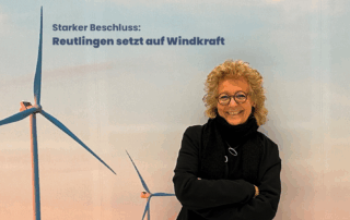 Beate Müller-Gemmeke steht lächelnd vor einem großen Hintergrundbild mit mehreren Windrädern im Abendlicht. Über ihr steht der Text: Starker Beschluss: Reutlingen setzt auf Windkraft. Sie trägt eine dunkle Jacke, Schal und Brille und hat die Arme entspannt verschränkt. Im Hintergrund ragen die Windräder in einen rosig-blauen Himmel.