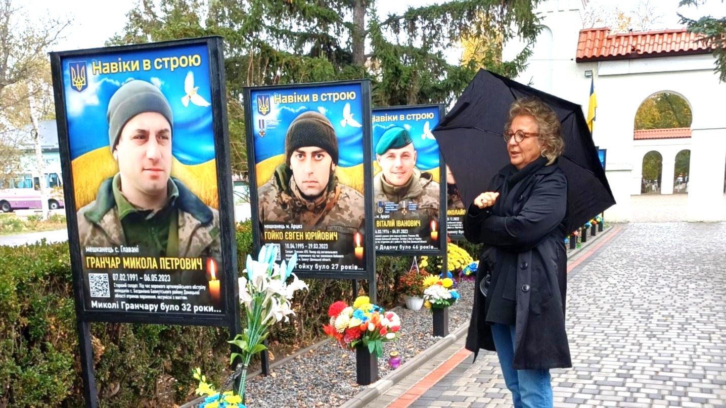 Beate Müller-Gemmeke steht mit schwarzem Mantel und aufgespanntem Regenschirm vor einer Gedenkstätte in Arzis. Mehrere große Porträttafeln gefallener ukrainischer Soldaten mit Flaggen und Blumen erinnern an junge Männer, die im Krieg getötet wurden. Die Szene wirkt still und würdevoll.