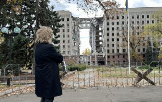 Beate Müller-Gemmeke steht vor einem schwer zerstörten Verwaltungsgebäude in Mykolajiw. Die ganze mittlere Gebäudefront ist herausgerissen, Beton hängt in großen Brocken herab. Herbstlaub liegt am Boden, davor ein Absperrgitter. Die Zerstörung zeigt die Folgen der russischen Angriffe.