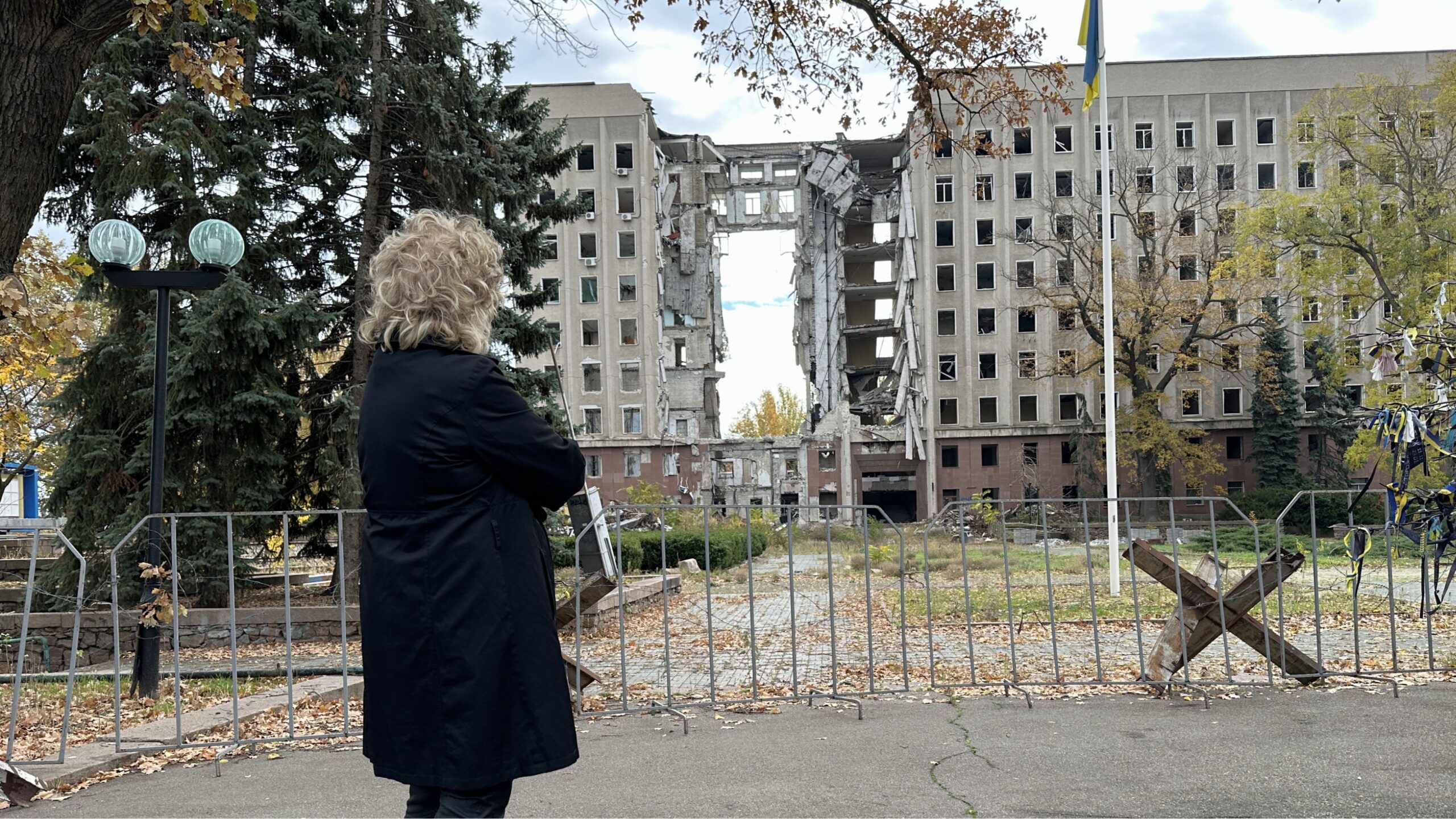 Beate Müller-Gemmeke steht vor einem schwer zerstörten Verwaltungsgebäude in Mykolajiw. Die ganze mittlere Gebäudefront ist herausgerissen, Beton hängt in großen Brocken herab. Herbstlaub liegt am Boden, davor ein Absperrgitter. Die Zerstörung zeigt die Folgen der russischen Angriffe.