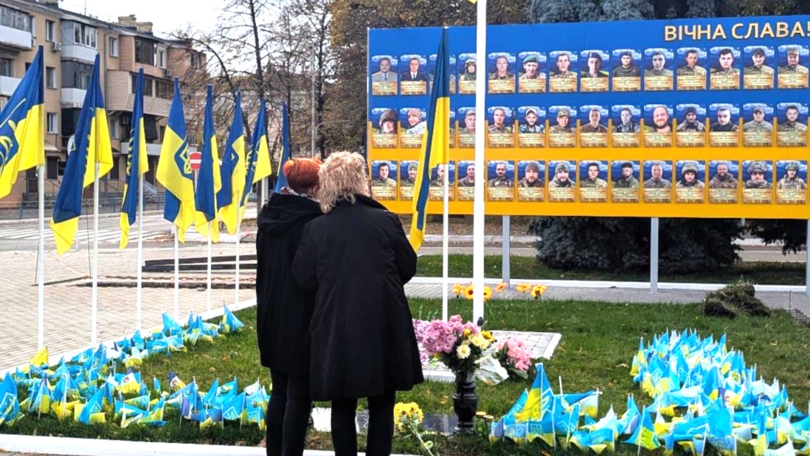 Beate Müller-Gemmeke und eine weitere Frau stehen vor einer großen Gedenkwand in Ismajil. Auf gelb-blauem Hintergrund sind zahlreiche Porträts gefallener Soldaten zu sehen. Umgeben ist die Wand von ukrainischen Fahnen und Blumen. Ein Ort des Gedenkens und des Verlusts.
