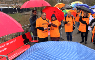 Beate Müller-Gemmeke und mehrere Personen stehen in orangefarbenen T-Shirts mit der Aufschrift „Stopp! Gewalt gegen Frauen“ im Regen zusammen. Viele halten bunte Regenschirme. Im Vordergrund spricht OB Thomas Keck, im Hintergrund versammeln sich weitere Menschen zur Aktion gegen Gewalt an Frauen.