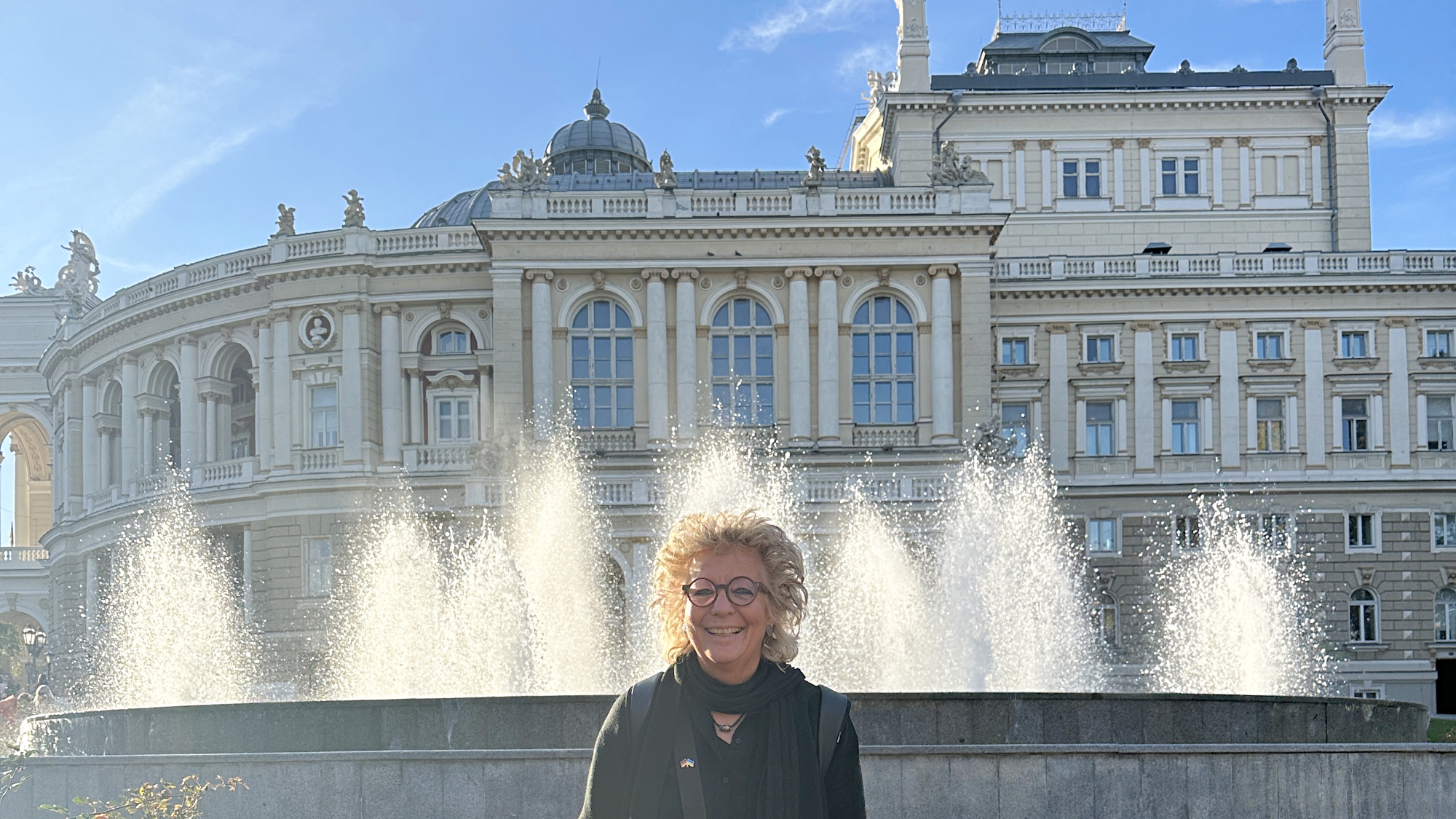Beate Müller-Gemmeke steht vor dem Opernhaus Odessa mit Sonnenschein, Wasserfontänen und klassischer Fassade im Hintergrund.