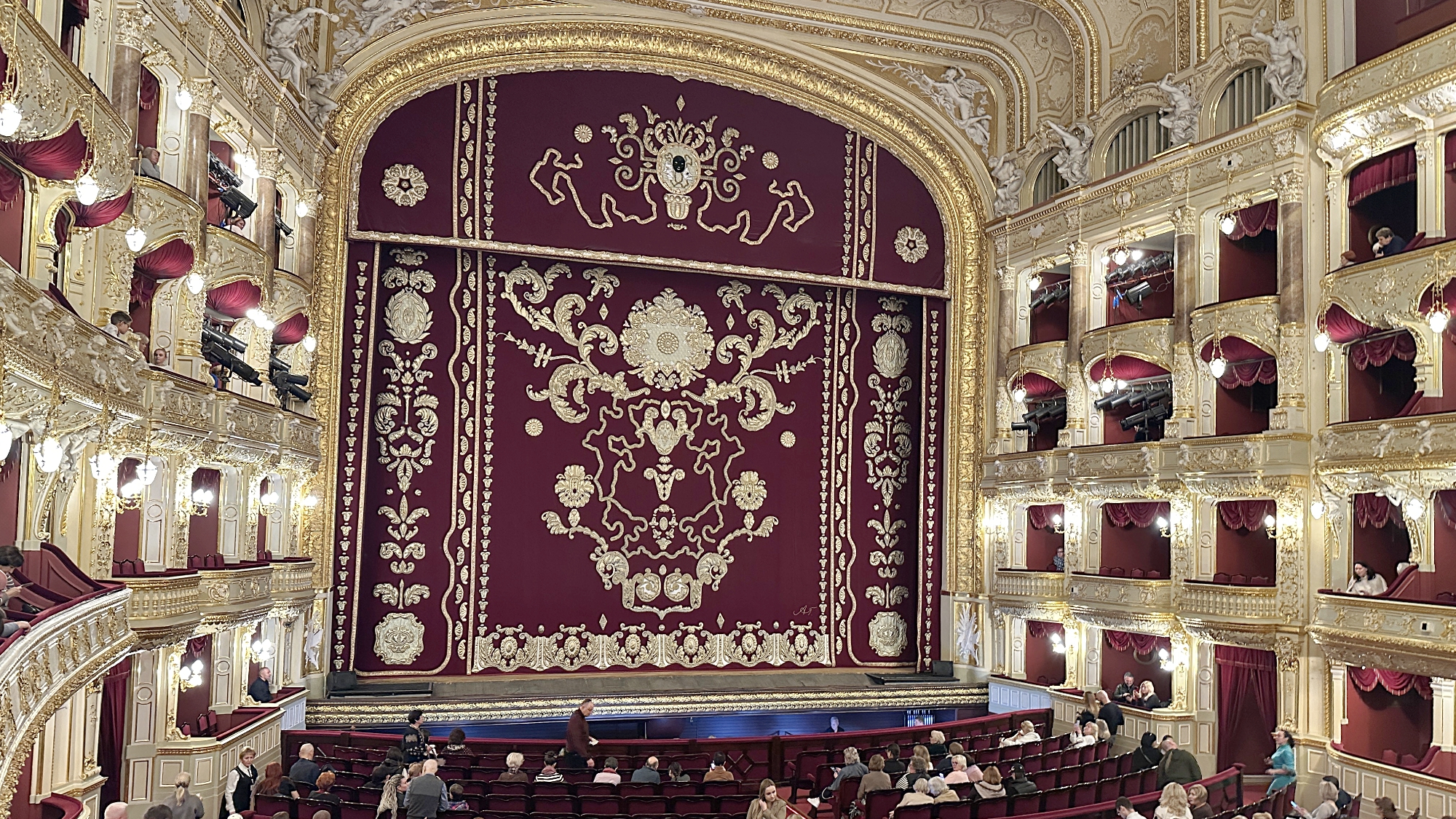 Blick in den Opernsaal von Odessa vor Beginn der Vorstellung – roter Samtvorhang mit goldenen Ornamenten, Publikum wartet gespannt.