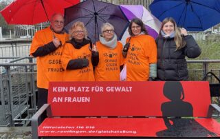 Auf dem Bild steht Beate Müller-Gemmeke zusammen mit vier weiteren Personen hinter der roten Bank „Kein Platz für Gewalt an Frauen“. Alle tragen orangefarbene T-Shirts mit der Aufschrift „Stopp! Gewalt gegen Frauen“ und halten bunte Regenschirme. Die Szene spielt vor der Stadthalle Reutlingen.