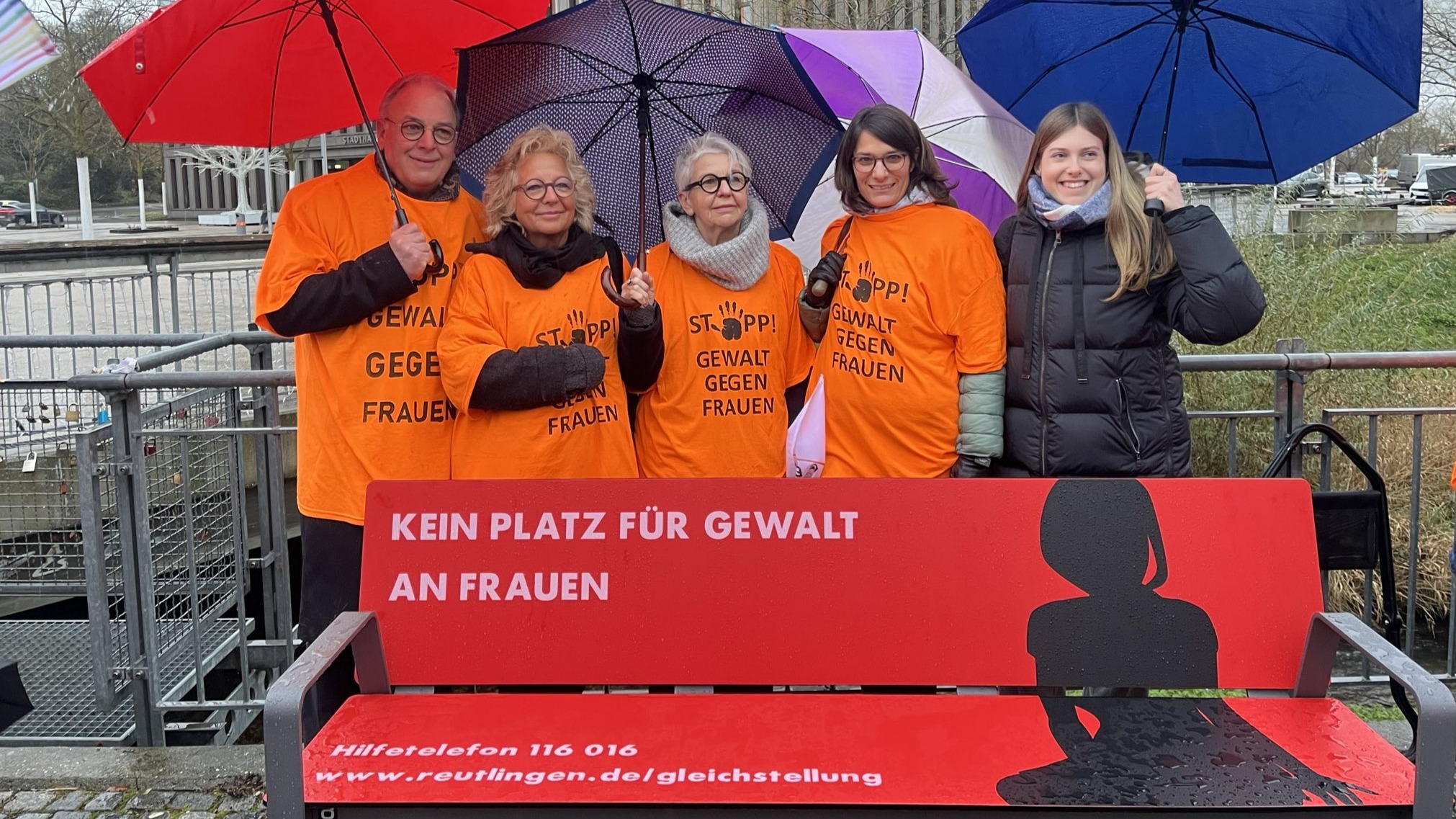 Auf dem Bild steht Beate Müller-Gemmeke zusammen mit vier weiteren Personen hinter der roten Bank „Kein Platz für Gewalt an Frauen“. Alle tragen orangefarbene T-Shirts mit der Aufschrift „Stopp! Gewalt gegen Frauen“ und halten bunte Regenschirme. Die Szene spielt vor der Stadthalle Reutlingen.