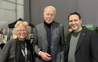 Beate Müller-Gemmeke steht in einer Messehalle der BDK Hannover neben Jürgen Trittin und Landtagskandidaten Kai Keune. Alle drei lächeln in die Kamera. Hinter ihnen sind Stände und weitere Delegierte zu sehen.