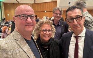 Beate Müller-Gemmeke steht in der Halle des grünen Landesparteitags in Ludwigsburg gemeinsam mit Michael Brecht und Cem Özdemir, im Hintergrund viele Teilnehmende.