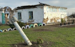 Beate Müller-Gemmeke dokumentiert ein zerstörtes Gebäude im Dorf Nowohryhoriwka. Im Vordergrund steckt ein Einschlagkörper einer russischen Rakete im Boden, dahinter steht ein schwer beschädigtes Haus mit eingeschlagenen Fenstern und einstürzenden Wänden.
