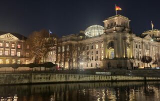 Das beleuchtete Reichstagsgebäude in Berlin bei Nacht, aufgenommen vom Spreeufer aus. Die Kuppel und die deutsche Flagge sind gut sichtbar, die Lichter spiegeln sich im Wasser.