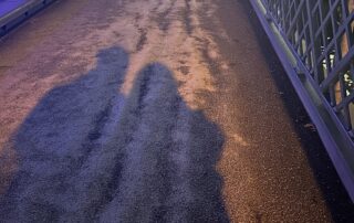 Zwei lange Schatten von Personen auf einem leicht nassen, farbig beleuchteten Boden. Die Umrisse sind deutlich, aber die Personen selbst außerhalb des Bildes.