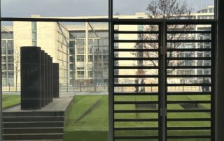 Blick aus einem Gebäude des Bundestags auf den Innenhof mit schwarzen Stelen und Grünflächen. Durch die Fensterfront sieht man moderne Regierungsgebäude und vorbeigehende Personen.