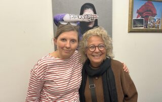 Beate Müller-Gemmeke steht Arm in Arm mit ihrer ehemaligen Kollegin Corinna Rüffer (MdB) im rot-weiß gestreiften Shirt. Beide lächeln in die Kamera. Im Hintergrund ein Foto einer Frau, die ihre Hand vor das Gesicht hält.