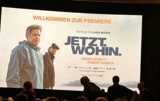 Eine große Kinoleinwand zeigt das Plakat des Films „JETZT. WOHIN. Meine Reise mit Robert Habeck“ von Lars Jessen. Links ist Robert Habeck im Windbreaker zu sehen, daneben Lars Jessen mit Kappe. Vor der Leinwand sitzen zahlreiche Menschen im abgedunkelten Saal. Oben steht in Orange „Willkommen zur Premiere“.