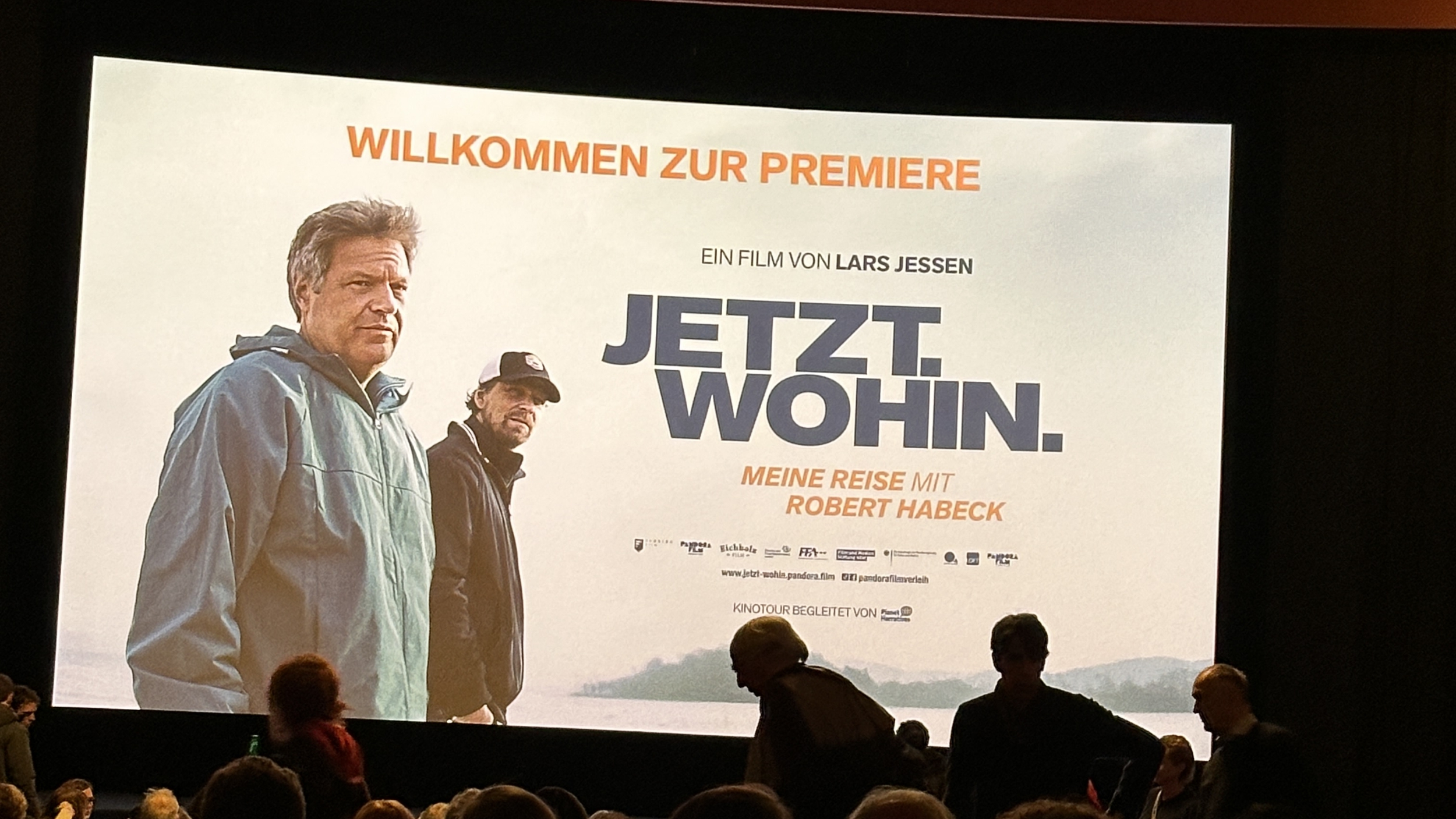 Eine große Kinoleinwand zeigt das Plakat des Films „JETZT. WOHIN. Meine Reise mit Robert Habeck“ von Lars Jessen. Links ist Robert Habeck im Windbreaker zu sehen, daneben Lars Jessen mit Kappe. Vor der Leinwand sitzen zahlreiche Menschen im abgedunkelten Saal. Oben steht in Orange „Willkommen zur Premiere“.