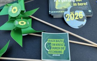Nahaufnahme von Materialien auf dem Grünen-Infostand: grüne Windräder aus Papier, Flyer und Aufkleber. Gut lesbar ist der Aufkleber mit dem Text „Mach Nazis ein Kreuz durch die Rechnung“.