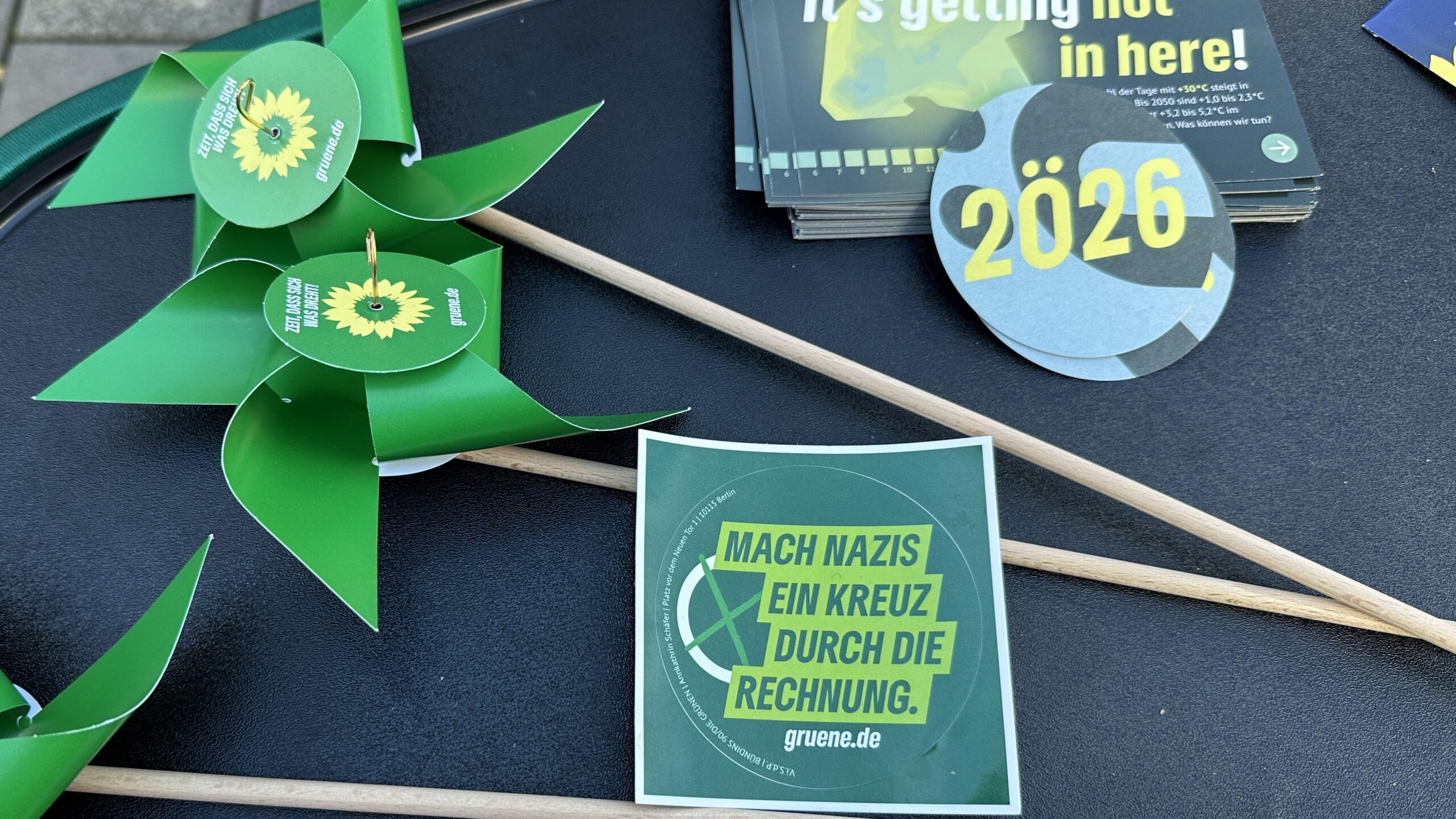 Nahaufnahme von Materialien auf dem Grünen-Infostand: grüne Windräder aus Papier, Flyer und Aufkleber. Gut lesbar ist der Aufkleber mit dem Text „Mach Nazis ein Kreuz durch die Rechnung“.