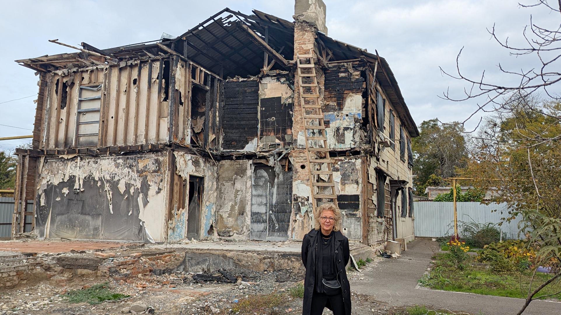 Beate Müller-Gemmeke steht vor einem ausgebrannten, teilweise eingestürzten Wohnhaus. Die Fassade ist schwarz, Wände fehlen, Holzträger hängen heraus. Das Bild zeigt die massiven Zerstörungen im Süden der Ukraine.