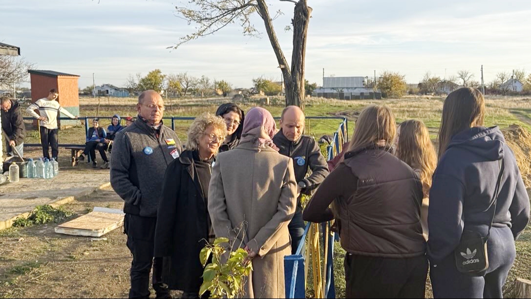 Beate Müller-Gemmeke spricht gemeinsam mit Mitgliedern der Delegation mit Jugendlichen aus Nowohryhoriwka. Die Gruppe steht auf dem Gelände des neu eingerichteten „Jugendtreff“, im Hintergrund einfache Gebäude und Felder. Es geht um Alltag, Rückkehr und Wiederaufbau.
