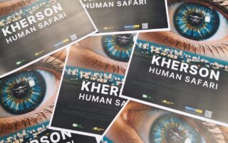 Mehrere ausgelegte Filmplakate mit dem Titel „Kherson: Human Safari“. Zu sehen ist ein Auge, in dessen Spiegelung eine Drohne erscheint, darunter der Schriftzug des Films. Die Plakate verweisen auf den Dokumentarfilm über den Krieg in der ukrainischen Stadt Cherson.