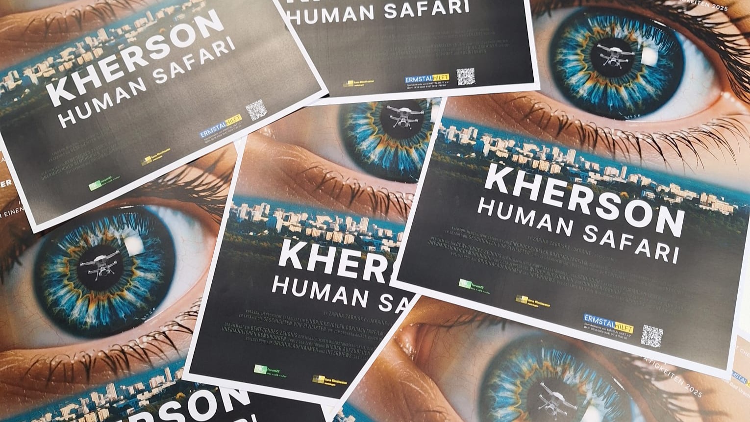 Mehrere ausgelegte Filmplakate mit dem Titel „Kherson: Human Safari“. Zu sehen ist ein Auge, in dessen Spiegelung eine Drohne erscheint, darunter der Schriftzug des Films. Die Plakate verweisen auf den Dokumentarfilm über den Krieg in der ukrainischen Stadt Cherson.