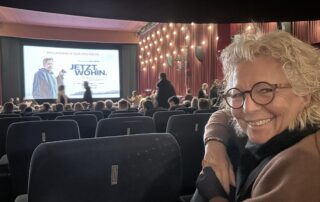Beate Müller-Gemmeke sitzt lächelnd in einem vollen Kinosaal und schaut in die Kamera. Im Hintergrund ist die beleuchtete Bühne zu sehen und auf der Leinwand das Plakat zum Film „JETZT. WOHIN.“. Die Stimmung ist erwartungsvoll und lebendig.