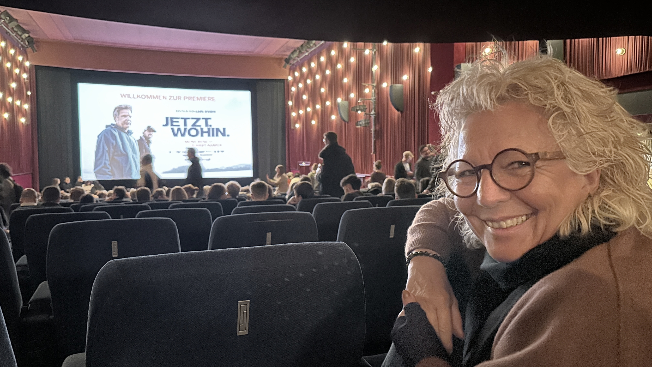 Beate Müller-Gemmeke sitzt lächelnd in einem vollen Kinosaal und schaut in die Kamera. Im Hintergrund ist die beleuchtete Bühne zu sehen und auf der Leinwand das Plakat zum Film „JETZT. WOHIN.“. Die Stimmung ist erwartungsvoll und lebendig.