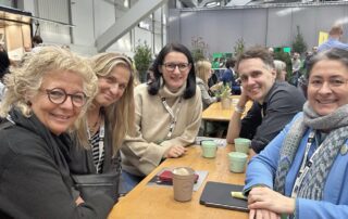 Beate Müller-Gemmeke sitzt mit vier (ehemalige) Kolleg:innen an einem Holztisch in der Pausenhalle der BDK Hannover. Sie lächeln, trinken Kaffee und wirken im Gespräch vertieft. Die Atmosphäre ist lebendig und freundlich.