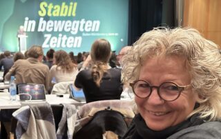 Beate Müller-Gemmeke sitzt in der Halle des Landesparteitags, im Hintergrund die Bühne mit dem Schriftzug Stabil in bewegten Zeiten und zahlreiche Delegierte.