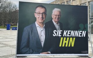 Großes grünes Wahlplakat mit Cem Özdemir und Winfried Kretschmann vor einem Gebäude, auf dem Plakat steht der Schriftzug Sie kennen ihn.