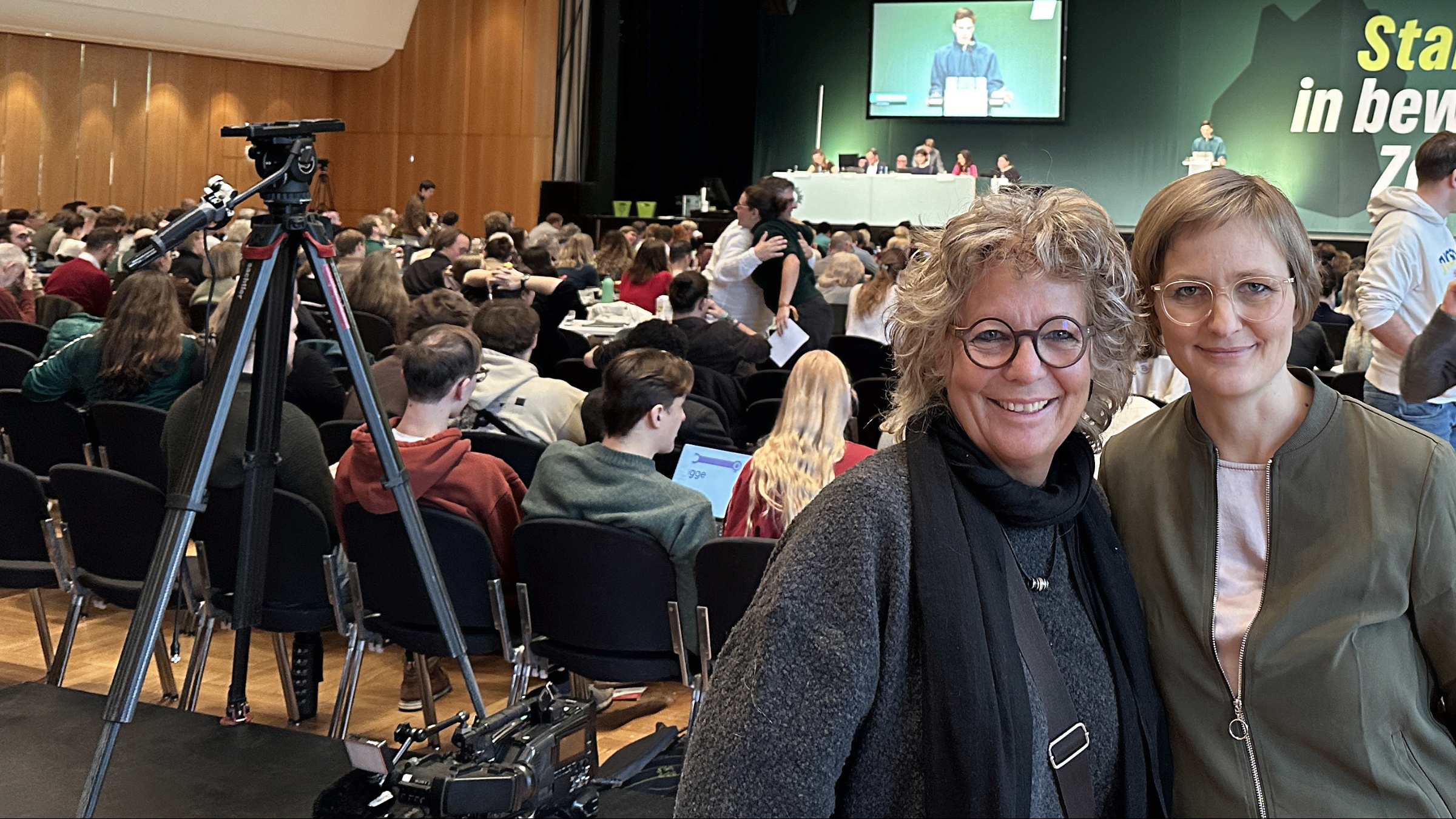 Beate Müller-Gemmeke steht gemeinsam mit Franziska Brantner in der Halle des Landesparteitags, im Hintergrund läuft die Debatte auf der Bühne.