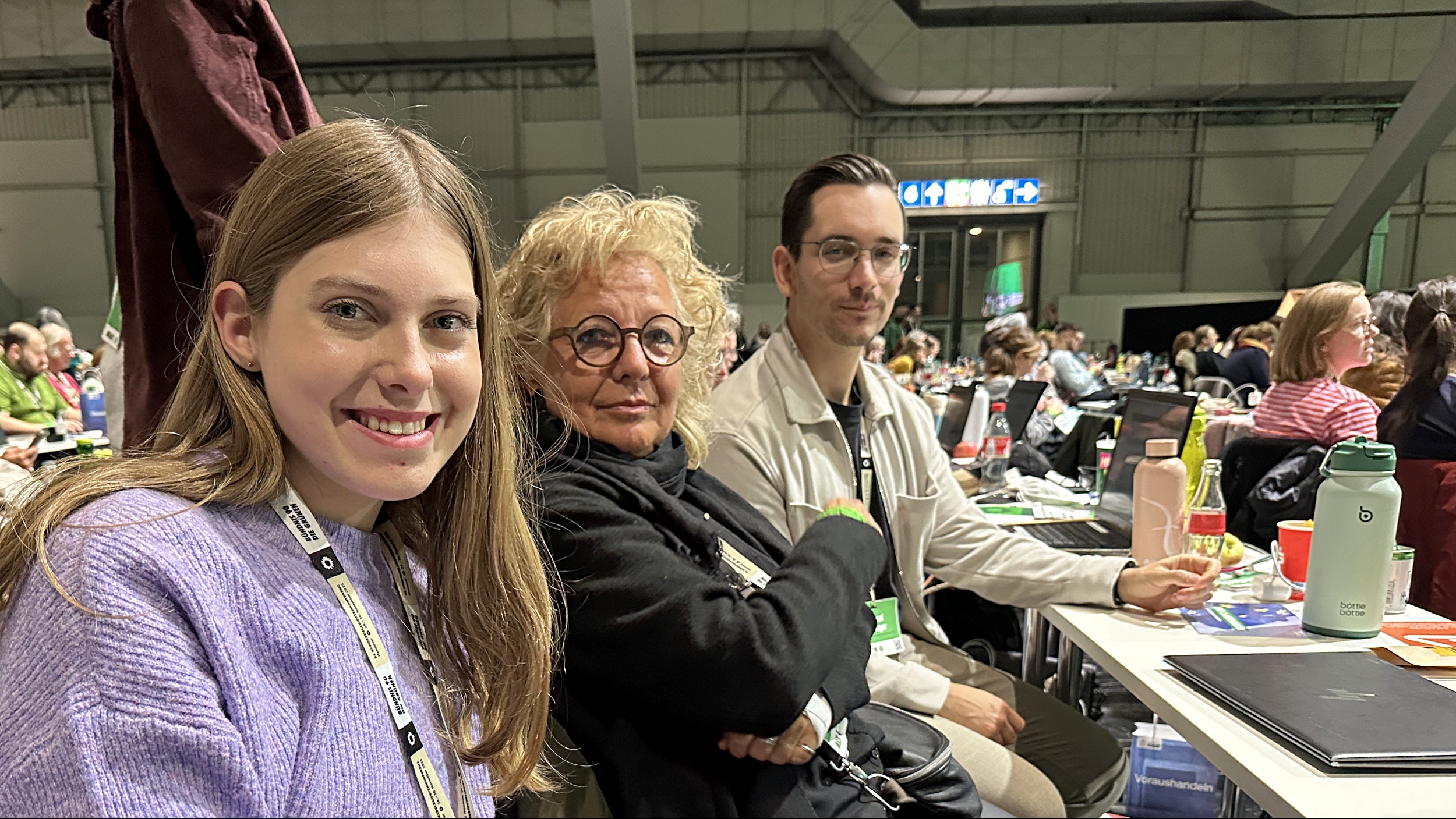 Beate Müller-Gemmeke sitzt im Plenumsbereich der BDK Hannover neben zwei jungen Delegierten. Alle drei lächeln, tragen Badges und sitzen an einem Tisch mit Unterlagen. Im Hintergrund ist der große Saal mit vielen Teilnehmenden zu sehen.