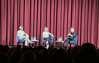 Auf der Kinobühne sitzen drei Personen auf Stühlen vor einem roten Vorhang: Lars Jessen, eine Moderatorin und Robert Habeck. Sie führen ein Gespräch nach der Filmvorführung. Auf kleinen Tischen stehen Wasserflaschen. Das Publikum schaut von unten auf die Bühne.