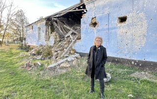 Beate Müller-Gemmeke steht vor einer komplett zerstörten Schule in Nowohryhoriwka. Die blaue Fassade ist von Einschüssen übersät, Teile des Gebäudes sind eingestürzt, Trümmer liegen auf dem Boden.