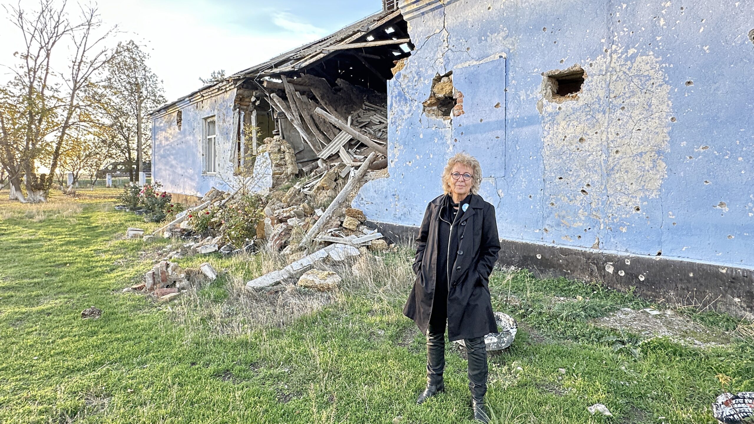 Beate Müller-Gemmeke steht vor einer komplett zerstörten Schule in Nowohryhoriwka. Die blaue Fassade ist von Einschüssen übersät, Teile des Gebäudes sind eingestürzt, Trümmer liegen auf dem Boden.