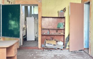 Innenraum eines zerstörten Klassenzimmers: umgestürzte Möbel, Schutt auf dem Boden, beschädigte Wände und offene Türen. Das Bild zeigt, wie russische Angriffe Bildungseinrichtungen verwüstet haben.
