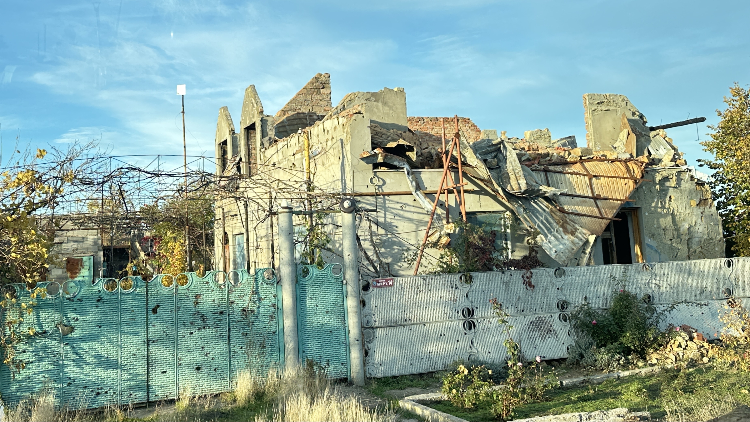 Blick auf ein zerstörtes Wohnhaus im ehemals besetzen Dorf Nowohryhoriwka in der Region Mykolajiw: eingestürztes Dach, freiliegende Wände und Einschusslöcher. Der Garten davor steht in starkem Kontrast zur massiven Kriegszerstörung.