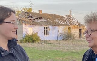 Beate Müller-Gemmeke spricht mit einer jungen Frau aus Nowohryhoriwka. Hinter den beiden steht ein zerstörtes Wohnhaus mit eingestürztem Dach. Die Begegnung zeigt persönliche Geschichten aus der Besatzungszeit.