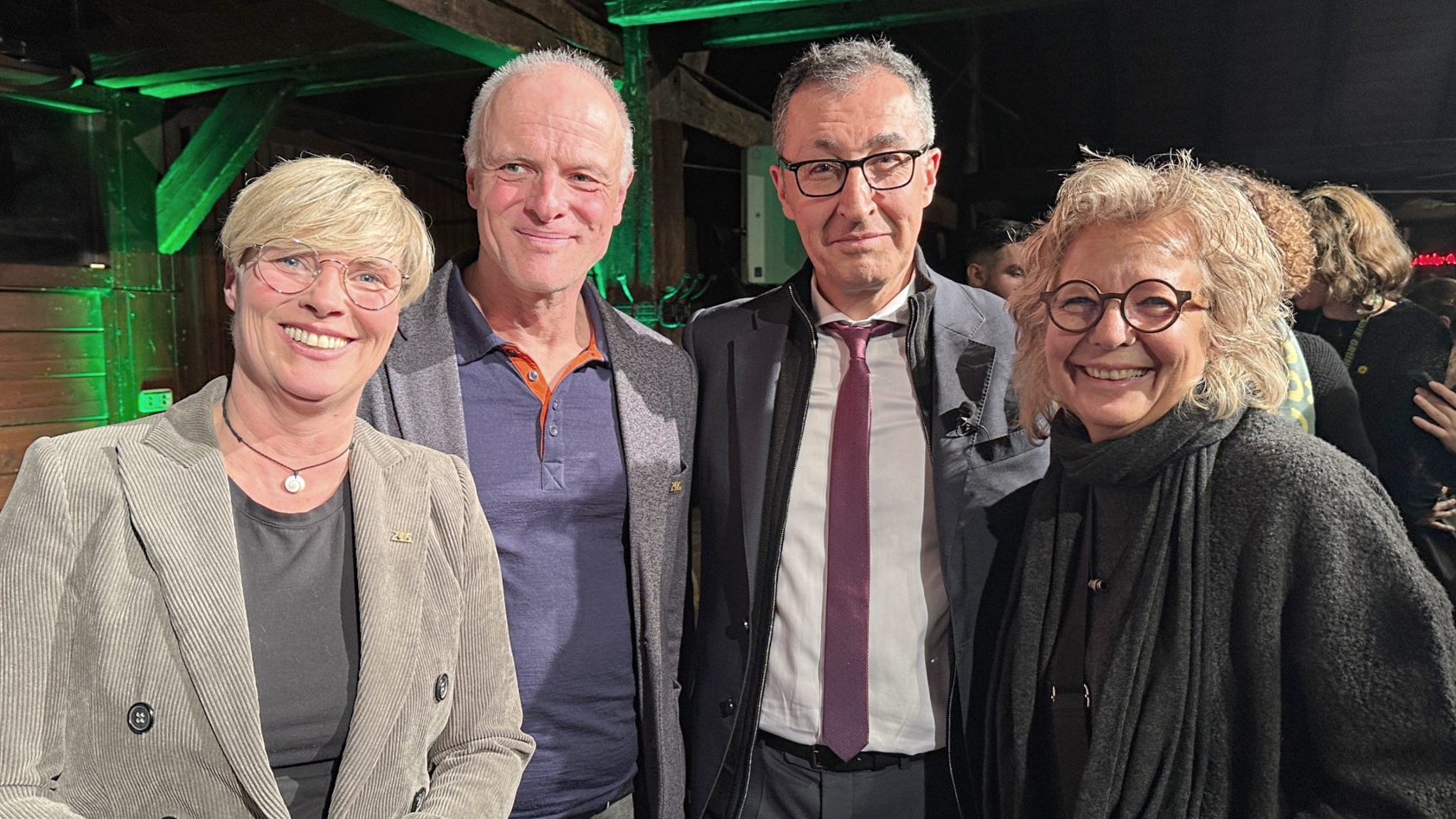 Vier Personen stehen eng nebeneinander in der historischen Festkelter in Metzingen und lächeln in die Kamera. In der Mitte steht Cem Özdemir, links und rechts von ihm stehen Cindy Holmberg, Thomas Poreski und Beate Müller-Gemmeke. Im Hintergrund sind Holzbalken der Festkelter zu sehen, die grün beleuchtet sind. Die Stimmung wirkt offen und herzlich.