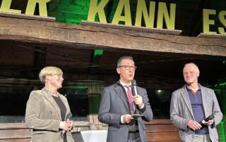 Cem Özdemir steht auf einer Bühne in der historischen Festkelter und spricht mit einem Mikrofon. Neben ihm stehen Cindy Holmberg und Thomas Poreski, ebenfalls mit Mikrofonen. Über der Bühne sind große grüne Buchstaben angebracht, die Teil einer Installation sind. Die Holzkonstruktion der Festkelter ist gut sichtbar, das Publikum befindet sich außerhalb des Bildes.