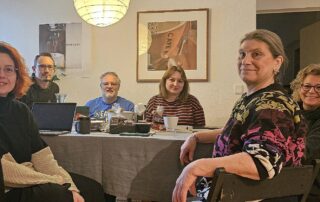 Gruppenfoto des Bundesvorstands von GewerkschaftsGrün in einem Innenraum: Sechs Personen sitzen an einem Tisch mit Laptops, Tassen und Unterlagen. Zu sehen sind (von links nach rechts) Yvonne Marchewitz, Klaudia Hanisch, Bernd Meffert und Dennis Kurzweil (Beisitzer:innen) sowie ganz links Sarah Gonschorek und ganz rechts Beate Müller-Gemmeke, die beiden Sprecherinnen von GewerkschaftsGrün. Die Gruppe blickt in die Kamera, die Atmosphäre wirkt konzentriert und kollegial.