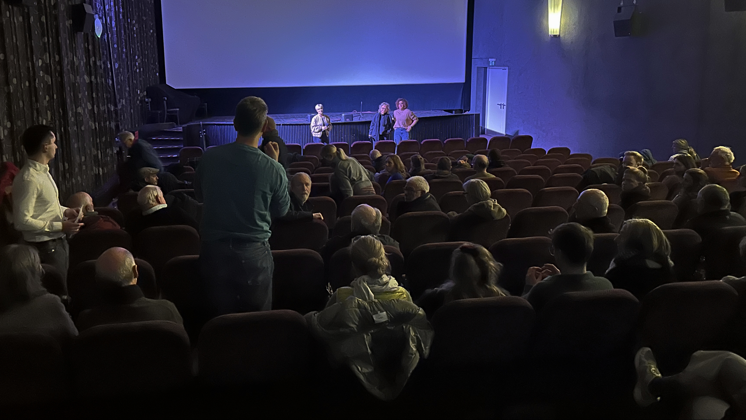Ein Kinobesucher steht im Mittelgang und stellt eine Frage. Im Vordergrund sitzen zahlreiche Besucherinnen und Besucher, im Hintergrund stehen Beate Müller-Gemmeke und Cindy Holmberg MdL vor der Leinwand.