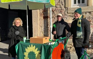 Beate Müller-Gemmeke steht lachend an einem grünen Infostand auf dem Marktplatz in Reutlingen. Neben ihr stehen Thomas Poreski und ein weiterer Unterstützer. Der Tisch ist mit einer grünen Fahne mit Sonnenblume bedeckt, im Hintergrund scheint die Sonne. Wahlkampfauftakt bei winterlichem, aber freundlichem Wetter.