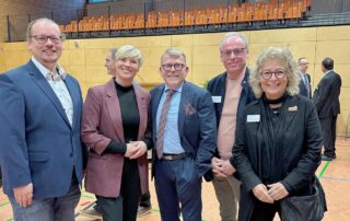 Alt-Text: Gruppenfoto beim Neujahrsempfang in Münsingen: In der Mitte Bürgermeister Mike Münzing. Links von ihm Cindy Holmberg, Landtagsabgeordnete und Kandidatin, sowie Beate Müller-Gemmeke. Daneben zwei grüne Gemeinderäte aus Münsingen. Alle stehen in einer Sporthalle und lächeln in die Kamera.